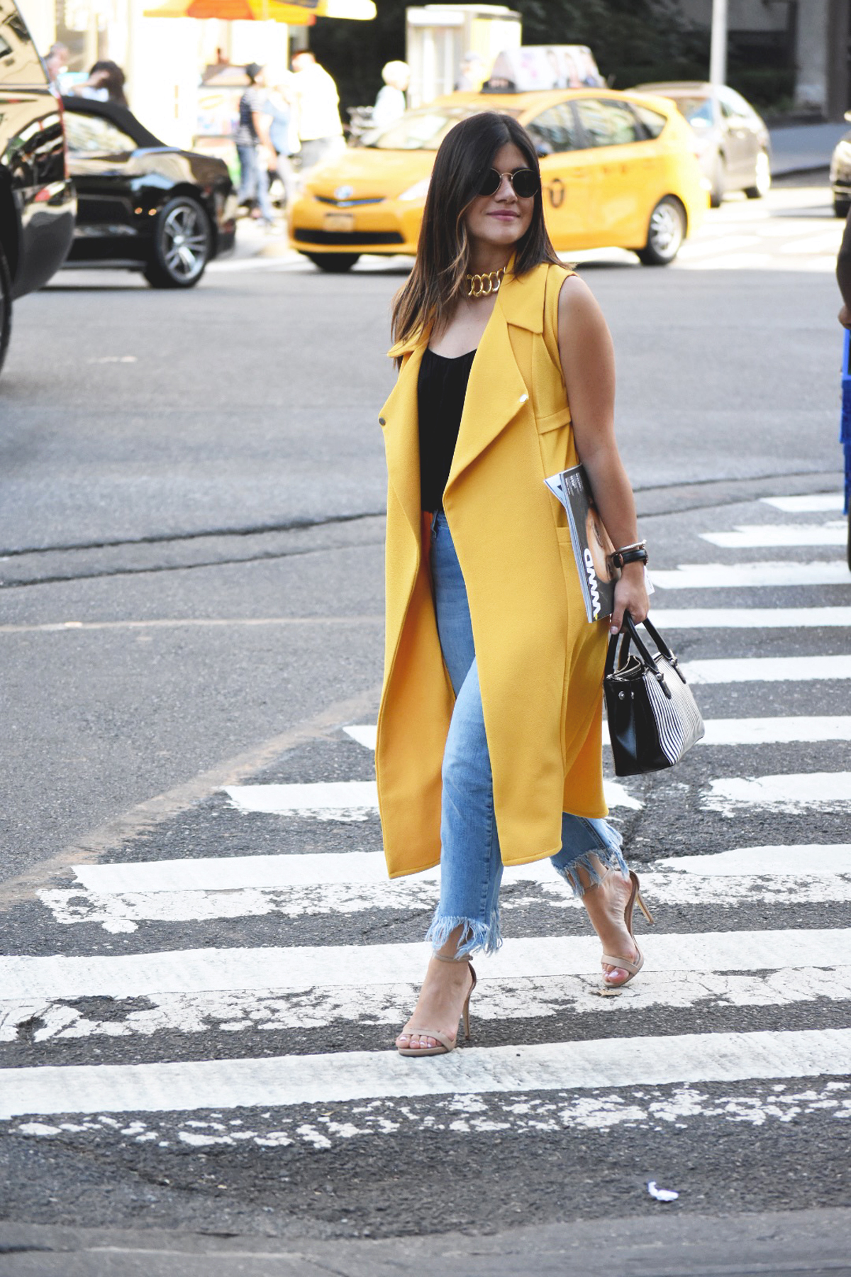 Carolina Hellal of Chic Talk wearing a Shein yellow maxi vest, Top shop fringed hem jeans, Rayban rounded sunglasses, Ralph Lauren striped bag, and Steve Madden beige strap sandals