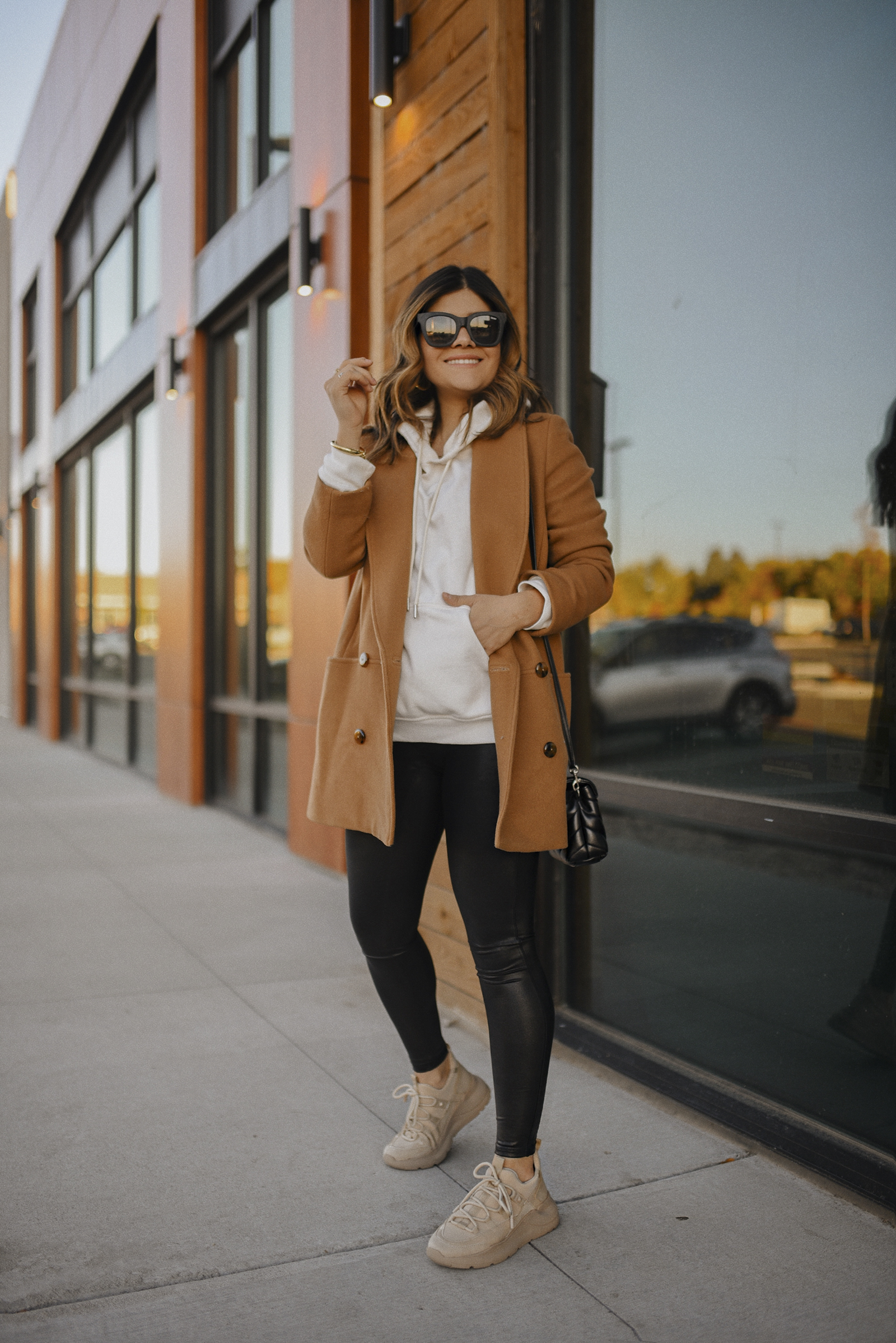 Carolina Hellal of Chic Talk wearing Sezane camel coat, Spanx faux leather leggings, H&M white hoodie , JLO Jennifer Lopez beige sneakers, quay Sunglasses and YSL crossbody bag